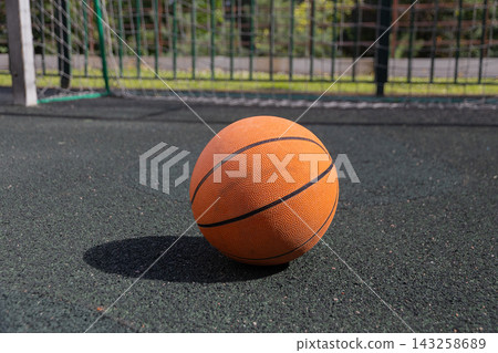 Close up of classic orange basketball on black rubber outdoor sports court surface. In the blurred background is soccer or handball goal net on sunny summer day. Street sports concept Close up of classic orange basketball on black rubber outdoor sports court surface. In the blurred background is soccer or handball goal net on sunny summer day. Street sports concept 143258689