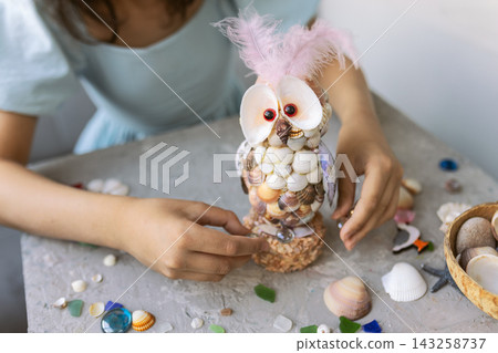 Close up of unrecognizable children's hands holding a creative owl craft made of various seashells and decorated with pink feathers. Handmade marine art project for kids with sea glass and shells on a Close up of unrecognizable children's hands holding a creative owl craft made of various seashells and decorated with pink feathers. Handmade marine art project for kids with sea glass and shells on a 143258737