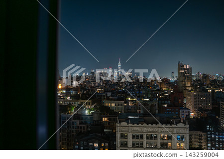 Wide panoramic view of New York City skyline at night with One World Trade Center glowing in Lower Manhattan. The urban cityscape is framed by nearby buildings and rooftops, highlighting illuminated Wide panoramic view of New York City skyline at night with One World Trade Center glowing in Lower Manhattan. The urban cityscape is framed by nearby buildings and rooftops, highlighting illuminated 143259004