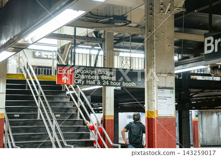 Exit sign directing to Brooklyn Bridge, Chambers Street and City Hall inside the subway station in Manhattan. Commuters move along the underground platform beneath fluorescent lighting. Exit sign directing to Brooklyn Bridge, Chambers Street and City Hall inside the subway station in Manhattan. Commuters move along the underground platform beneath fluorescent lighting. 143259017