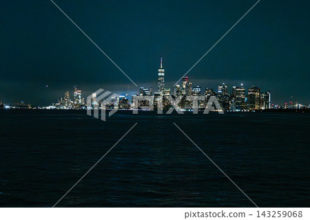 Lower Manhattan skyline at night with One World Trade Center illuminated above the city lights across the river. Iconic New York City cityscape featuring skyscrapers, dark water, and glowing downtown Lower Manhattan skyline at night with One World Trade Center illuminated above the city lights across the river. Iconic New York City cityscape featuring skyscrapers, dark water, and glowing downtown 143259068