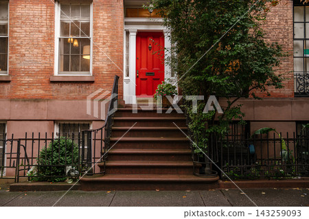 Bright red front door with brownstone stairs and black iron railing on a historic New York City brick townhouse. Traditional Manhattan residential architecture with sidewalk, greenery, and detailed Bright red front door with brownstone stairs and black iron railing on a historic New York City brick townhouse. Traditional Manhattan residential architecture with sidewalk, greenery, and detailed 143259093
