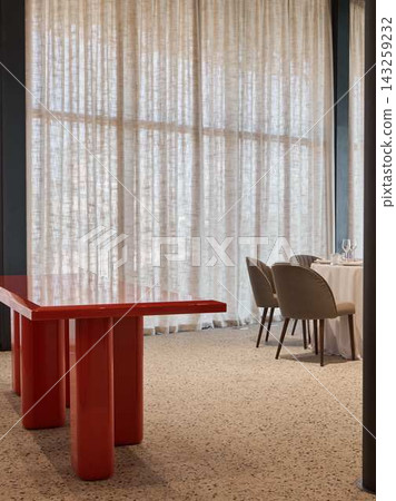 Red glossy dining table surrounded by plush beige chairs beside sheer curtains in elegant restaurant interior with soft daylight and warm inviting atmosphere Red glossy dining table surrounded by plush beige chairs beside sheer curtains in elegant restaurant interior with soft daylight and warm inviting atmosphere 143259232