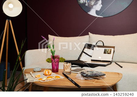 Living room scene with beige sofa, tripod wooden floor lamp, purple wall, round coffee table, open magazine, pink glass vase, orange peels on newspaper, desk accessories Living room scene with beige sofa, tripod wooden floor lamp, purple wall, round coffee table, open magazine, pink glass vase, orange peels on newspaper, desk accessories 143259240