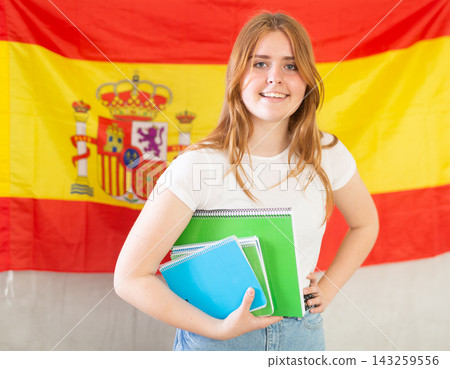Happy smiling teen girl holding spiral notepads under her arm against of national flag of Spain Happy smiling teen girl holding spiral notepads under her arm against of national flag of Spain 143259556