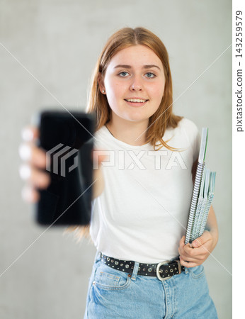 Smiling teenage girl, student with notebooks under her arm and mobile phone with blank black screen at gray wall background. Free copy space for advertisement Smiling teenage girl, student with notebooks under her arm and mobile phone with blank black screen at gray wall background. Free copy space for advertisement 143259579