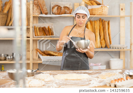Young female baker whips ingredients in metal bowl Young female baker whips ingredients in metal bowl 143259635