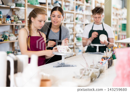 Young woman teaches teenage boy and teenage girl to paint ceramic cup 143259680