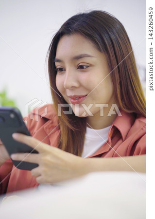 Asian woman texting on smartphone sitting relaxed in living room Asian woman texting on smartphone sitting relaxed in living room 143260459