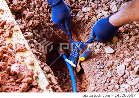 Worker fix blue water pipe in hole dug in soil with tools in hand. 143260597