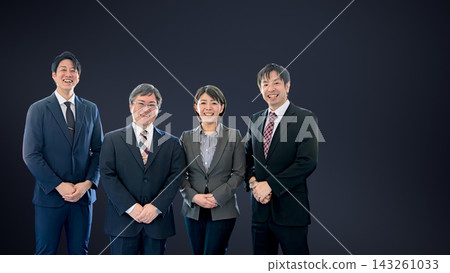 Four smiling businesspeople in suits stand side-by-side against a dark background, with a blank space on the right. Four smiling businesspeople in suits stand side-by-side against a dark background, with a blank space on the right. 143261033