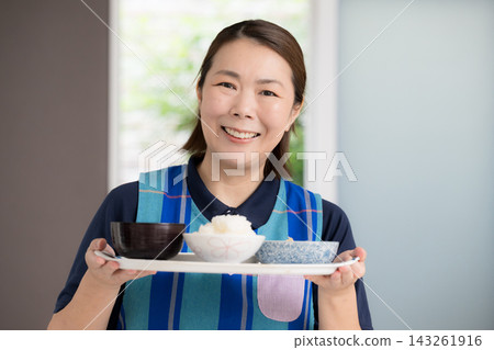 一位微笑的照護員正在準備餐點；一位家庭幫手提供餐食，這是家庭照護和家事服務的一部分；畫面直視鏡頭。 143261916