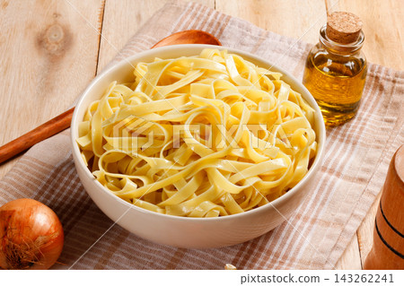 Prepared fettuccine pasta served in a bowl on a rustic cloth and wooden surface, with olive oil, garlic, tomato, and onion. 143262241