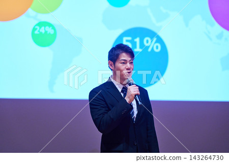 A Japanese man gives a presentation with a microphone in front of a screen. A Japanese man gives a presentation with a microphone in front of a screen. 143264730