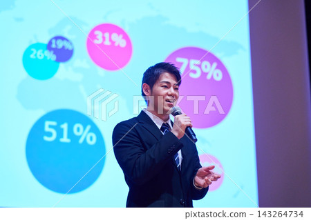 A Japanese man gives a presentation with a microphone in front of a screen. A Japanese man gives a presentation with a microphone in front of a screen. 143264734