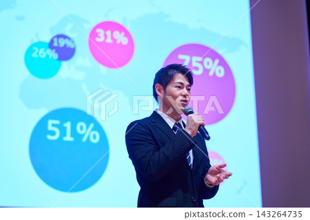 A Japanese man gives a presentation with a microphone in front of a screen. A Japanese man gives a presentation with a microphone in front of a screen. 143264735
