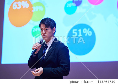 A Japanese man gives a presentation with a microphone in front of a screen. A Japanese man gives a presentation with a microphone in front of a screen. 143264739