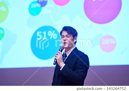 A Japanese man gives a presentation with a microphone in front of a screen. 143264752