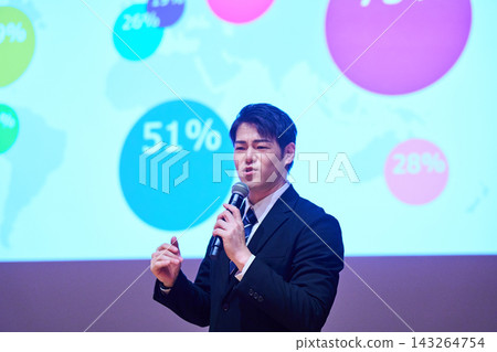 A Japanese man gives a presentation with a microphone in front of a screen. 143264754