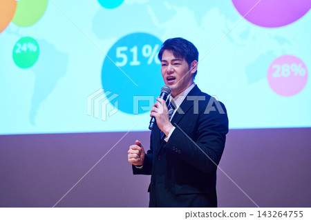 A Japanese man gives a presentation with a microphone in front of a screen. 143264755