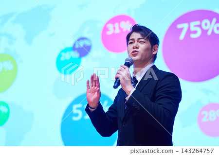 A Japanese man gives a presentation with a microphone in front of a screen. 143264757