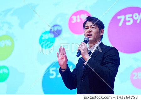 A Japanese man gives a presentation with a microphone in front of a screen. 143264758
