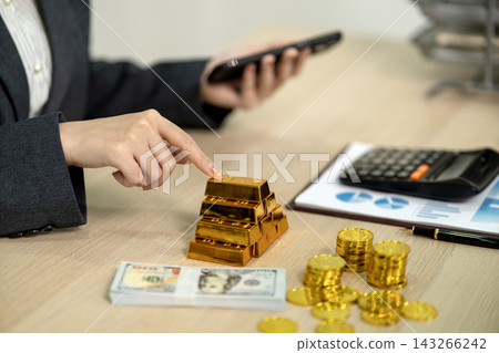 Businesswoman Touching Stacked Gold Bars While Using Smartphone at Office Desk with Cash and Coins Businesswoman Touching Stacked Gold Bars While Using Smartphone at Office Desk with Cash and Coins 143266242