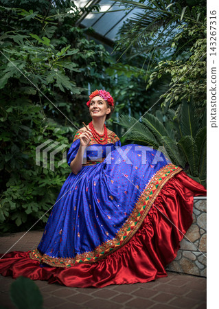 Smiling young girl in Frida Kahlo style dress sitting in a tropical greenhouse Smiling young girl in Frida Kahlo style dress sitting in a tropical greenhouse 143267316