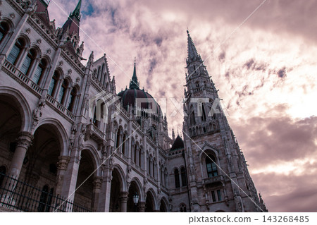 Hungarian parliament building in Budapest seen from a low angle against a dramatic cloudy sky, highlighting its ornate gothic revival dome, spires, turrets and detailed stone façade 143268485