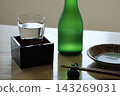冰鎮清酒倒入裝有枡（木製清酒杯）的玻璃杯中，旁邊是日式盤子、一瓶清酒和一個日式食品櫃檯。 143269031