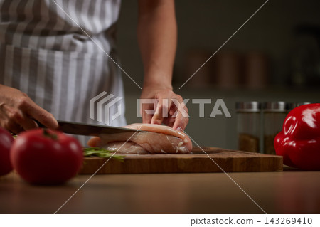 Chef preparing fresh raw chicken breast on wood cutting board, holding a sharp knife and slicing poultry meat Chef preparing fresh raw chicken breast on wood cutting board, holding a sharp knife and slicing poultry meat 143269410