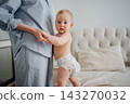 Cute baby holding mothers hand standing on bed in diaper 143270032