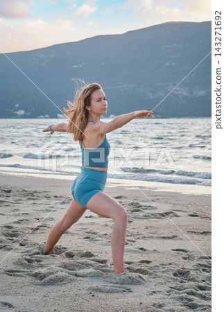 A woman stands on beach sand in Virabhadrasana 2 yoga, Warrior 2 Pose, with a bent front knee and arms extended sideways as she gazes along her forward hand 143271692