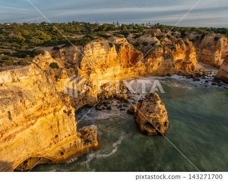 Drone view of Praia da Marinha in Algarve, Portugal, with golden limestone cliffs, a small sandy cove and an offshore sea stack in soft sunrise light. 143271700