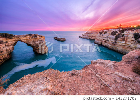 Dramatic sunset view of Arco de Albandeira at Praia de Albandeira in Algarve, Portugal, with pink sky, limestone sea arch, Atlantic water, and coastal shrubland above the cliff top. Dramatic sunset view of Arco de Albandeira at Praia de Albandeira in Algarve, Portugal, with pink sky, limestone sea arch, Atlantic water, and coastal shrubland above the cliff top. 143271702