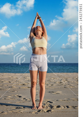 A woman looks upward in a front view of Urdhva Hastasana yoga, Upward Salute, extending her arms and joined palms overhead against a blue coastal sky A woman looks upward in a front view of Urdhva Hastasana yoga, Upward Salute, extending her arms and joined palms overhead against a blue coastal sky 143273931