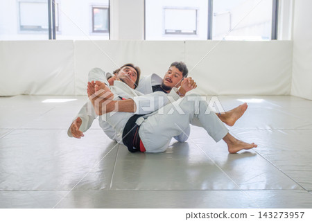 Two Brazilian jiu jitsu practitioners work on a collar choke from back control, with the attacking athlete holding the lapel and placing a leg across the body Two Brazilian jiu jitsu practitioners work on a collar choke from back control, with the attacking athlete holding the lapel and placing a leg across the body 143273957