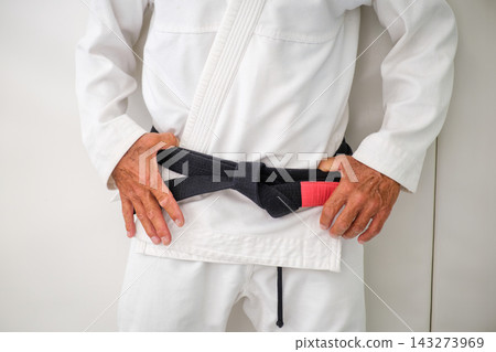Close up of a tied Brazilian jiu jitsu black belt with a red rank bar over a white gi, with the hands of the practitioner beside the knot 143273969