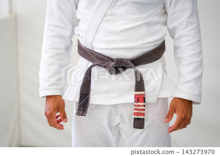 Close up of a worn Brazilian jiu jitsu brown belt tied over a white gi, with rank stripes on one end and the hands of the practitioner at the sides Close up of a worn Brazilian jiu jitsu brown belt tied over a white gi, with rank stripes on one end and the hands of the practitioner at the sides 143273970