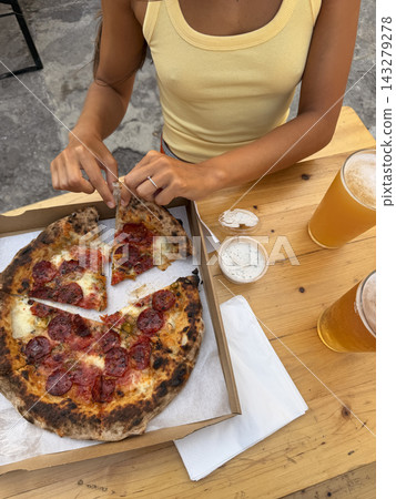 Woman in yellow ribbed tank top sharing pepperoni pizza at outdoor table with beer and dipping sauces, casual dining scene with hands reaching for slice, rustic wood setting and relaxed meal Woman in yellow ribbed tank top sharing pepperoni pizza at outdoor table with beer and dipping sauces, casual dining scene with hands reaching for slice, rustic wood setting and relaxed meal 143279278