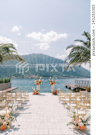 Elegant seaside wedding ceremony setup with chairs, floral arrangements, and mountain view Elegant seaside wedding ceremony setup with chairs, floral arrangements, and mountain view 143283305