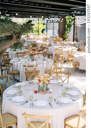 Elegant outdoor wedding reception table setting with floral centerpieces, round linen covered tables, wooden chairs, glassware, and warm string lights in bright garden venue Elegant outdoor wedding reception table setting with floral centerpieces, round linen covered tables, wooden chairs, glassware, and warm string lights in bright garden venue 143283587