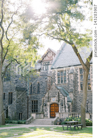 Historic stone manor with arched wooden entrance, tall windows, steep rooflines, and mature trees surrounding bright courtyard in soft morning sunlight, showing classic architecture and peaceful Historic stone manor with arched wooden entrance, tall windows, steep rooflines, and mature trees surrounding bright courtyard in soft morning sunlight, showing classic architecture and peaceful 143283907