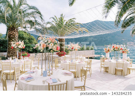 Elegant seaside wedding reception tables with flowers under palm trees beside mountain bay Elegant seaside wedding reception tables with flowers under palm trees beside mountain bay 143284235