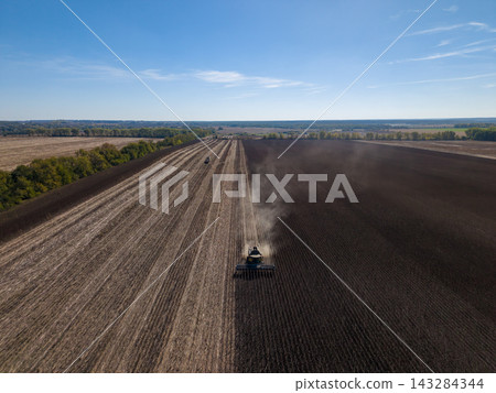Agricultural machines work across broad field, with no visible sunflower crop. Two tractors raise dust over parallel rows beneath open blue sky, showing expansive farmland prepared for cultivation. Agricultural machines work across broad field, with no visible sunflower crop. Two tractors raise dust over parallel rows beneath open blue sky, showing expansive farmland prepared for cultivation. 143284344