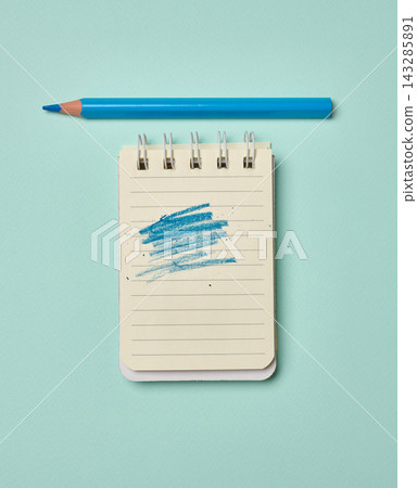 Spiral-bound lined notepad with a blue scribbled stroke and a blue pencil on a turquoise background. Spiral-bound lined notepad with a blue scribbled stroke and a blue pencil on a turquoise background. 143285891