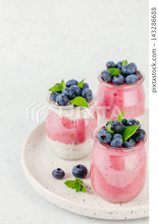 Three glass jars filled with layered yogurt dessert topped with fresh blueberries and mint leaves on a round plate against a light background. copy space Three glass jars filled with layered yogurt dessert topped with fresh blueberries and mint leaves on a round plate against a light background. copy space 143286688