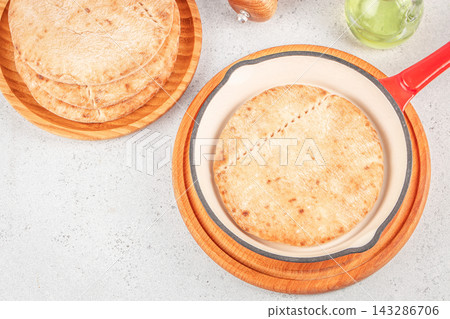 Top view of round flatbreads on wooden plates beside a red handled baking dish and green glassware on a light kitchen surface Top view of round flatbreads on wooden plates beside a red handled baking dish and green glassware on a light kitchen surface 143286706