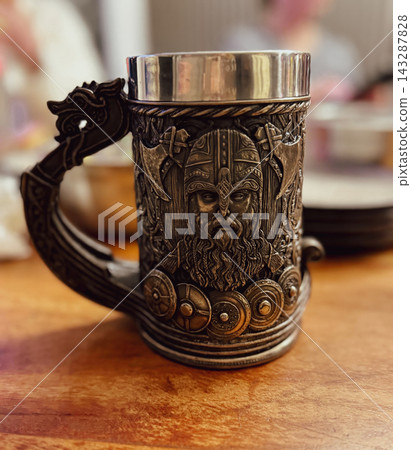 Ornate metal beer mug featuring a detailed Viking warrior in a horned helmet, shields and elaborate Norse-inspired decorations, photographed on a wooden table with a softly blurred background 143287828