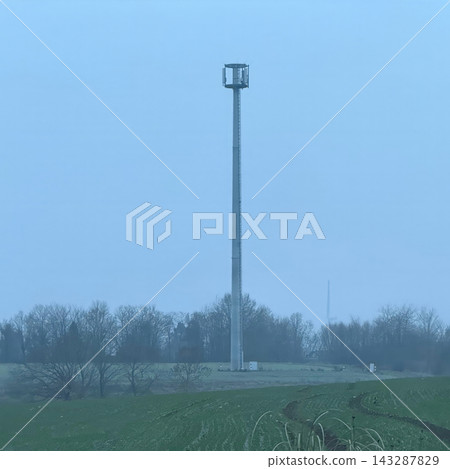 Tall cellular communication tower standing in a misty rural landscape with green agricultural fields and leafless trees on a winter morning Tall cellular communication tower standing in a misty rural landscape with green agricultural fields and leafless trees on a winter morning 143287829